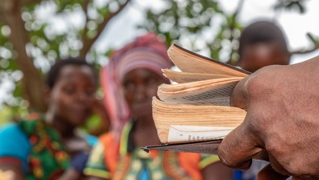 A close-up of a hand holding a book with blurred villagers in the background.