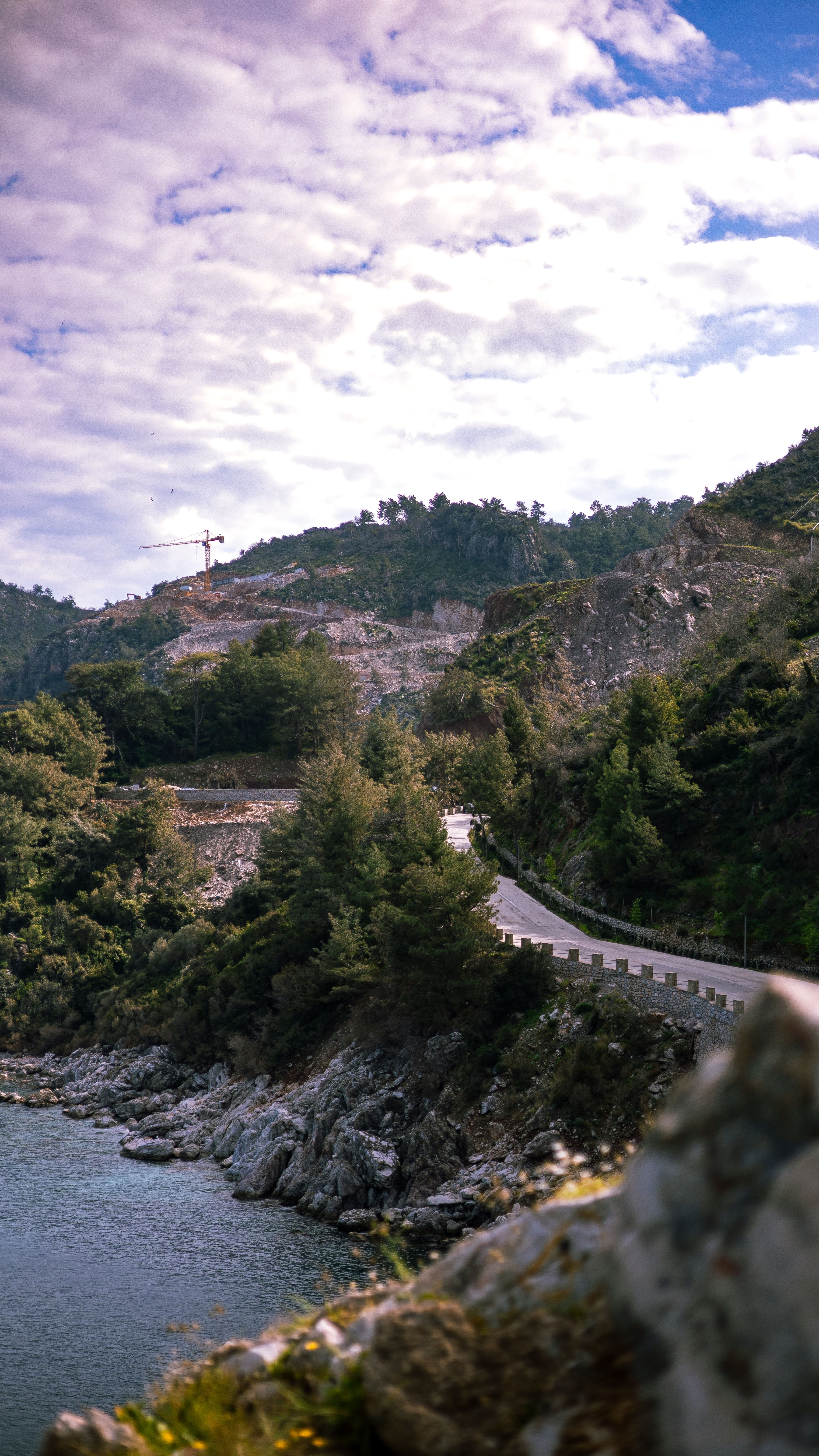 A winding road along a rocky coastline under a partly cloudy blue sky, surrounded by lush greenery.