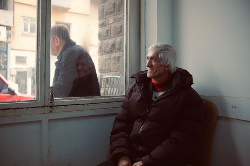 Free Elderly man warmly dressed sits by a window with a thoughtful expression, observing the outside world. Stock Photo