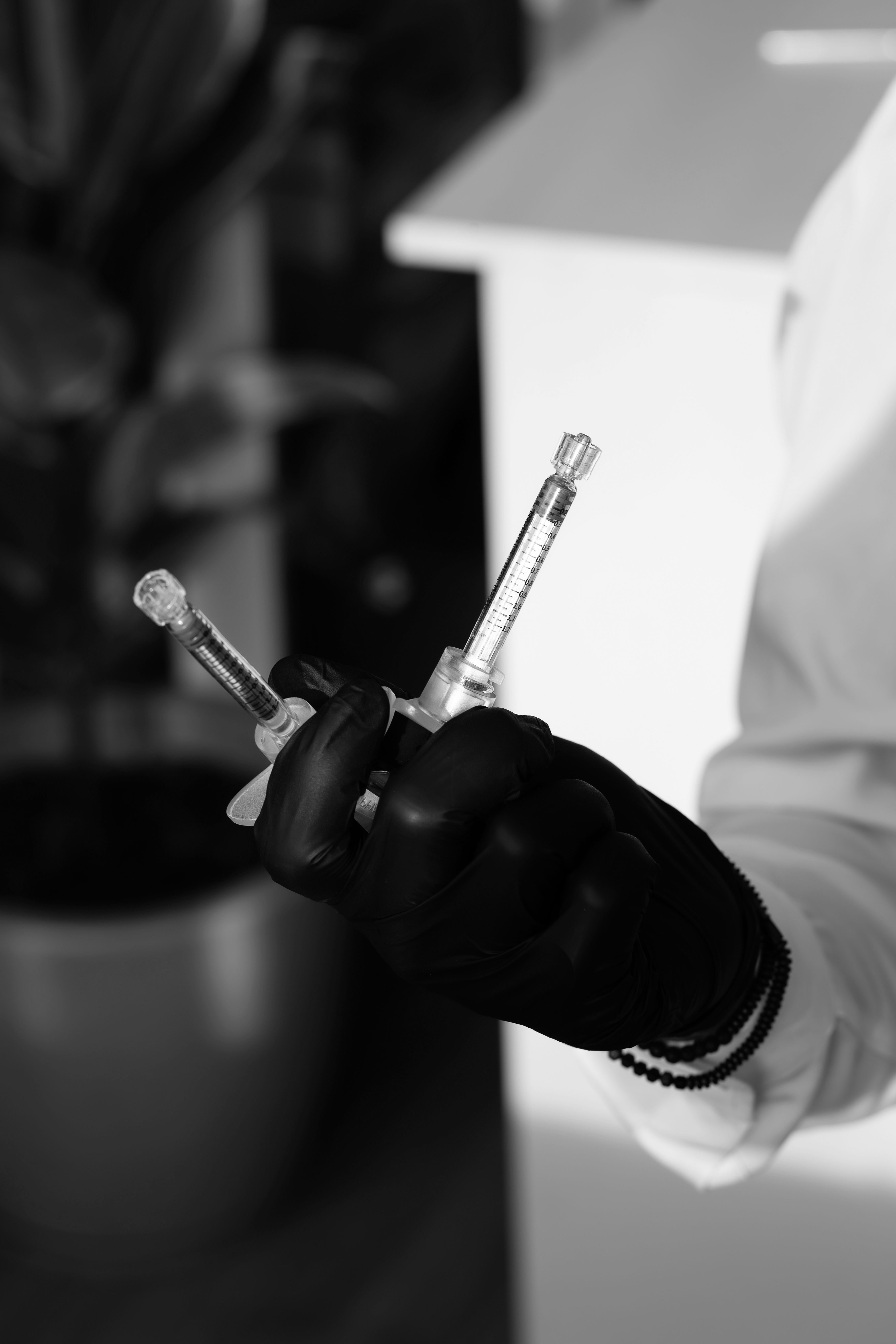 Monochrome photo of two syringes held by a person wearing black gloves, indoors.