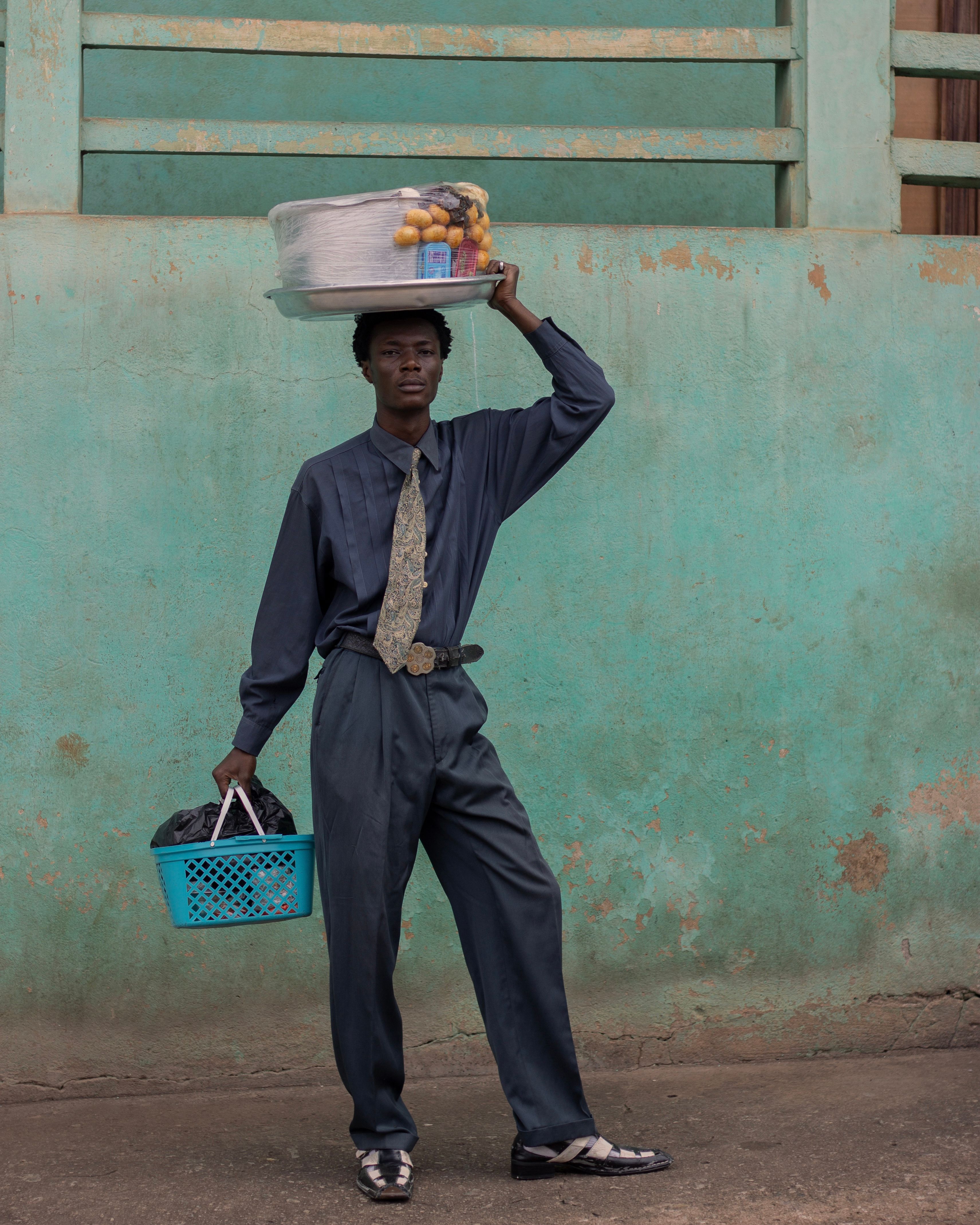 A vendor in blue attire balances a fruit tray on his head and holds a basket, standing against a teal wall.