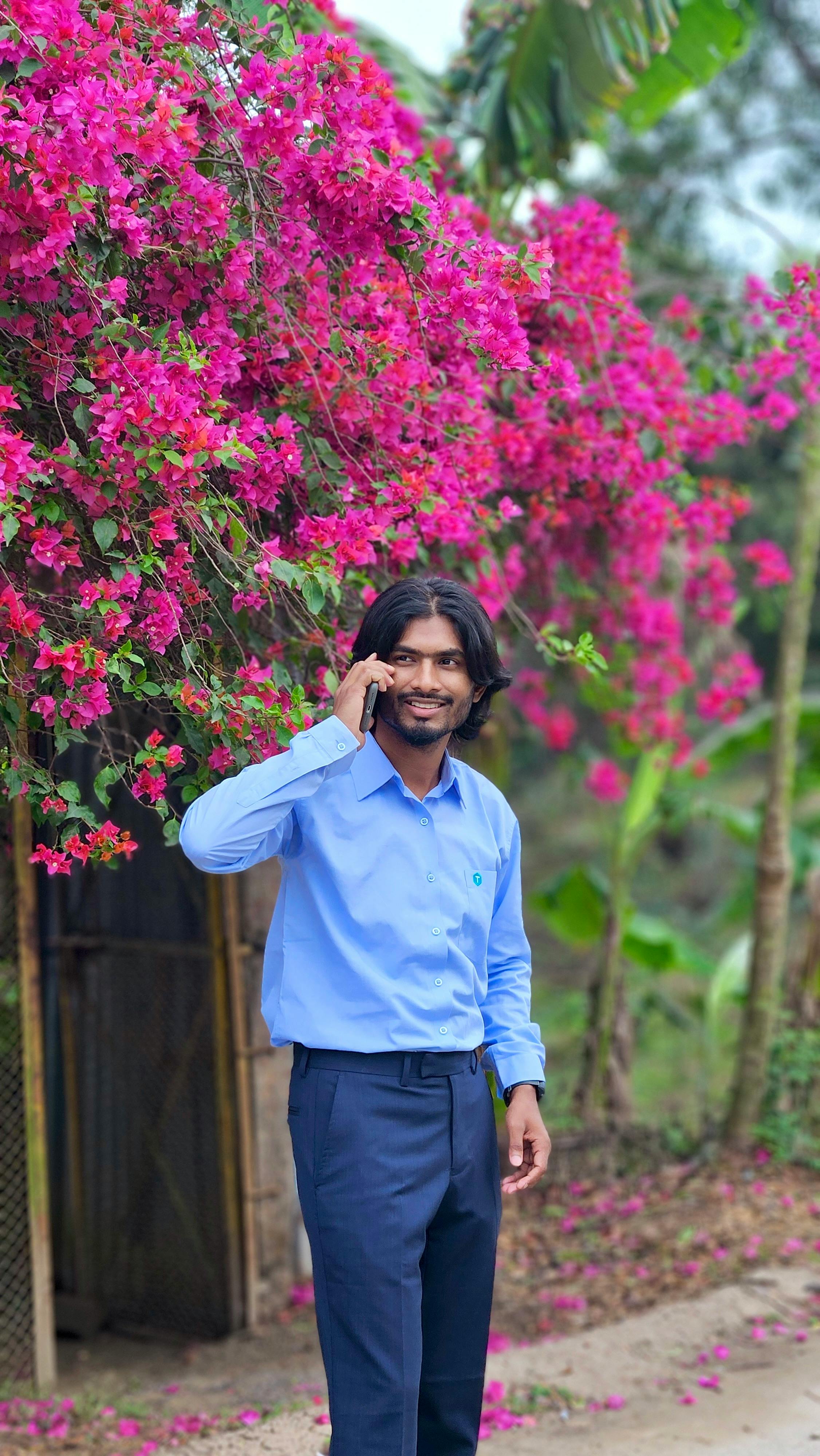 A man in a blue shirt talking on a phone under vibrant pink bougainvillea outdoors.