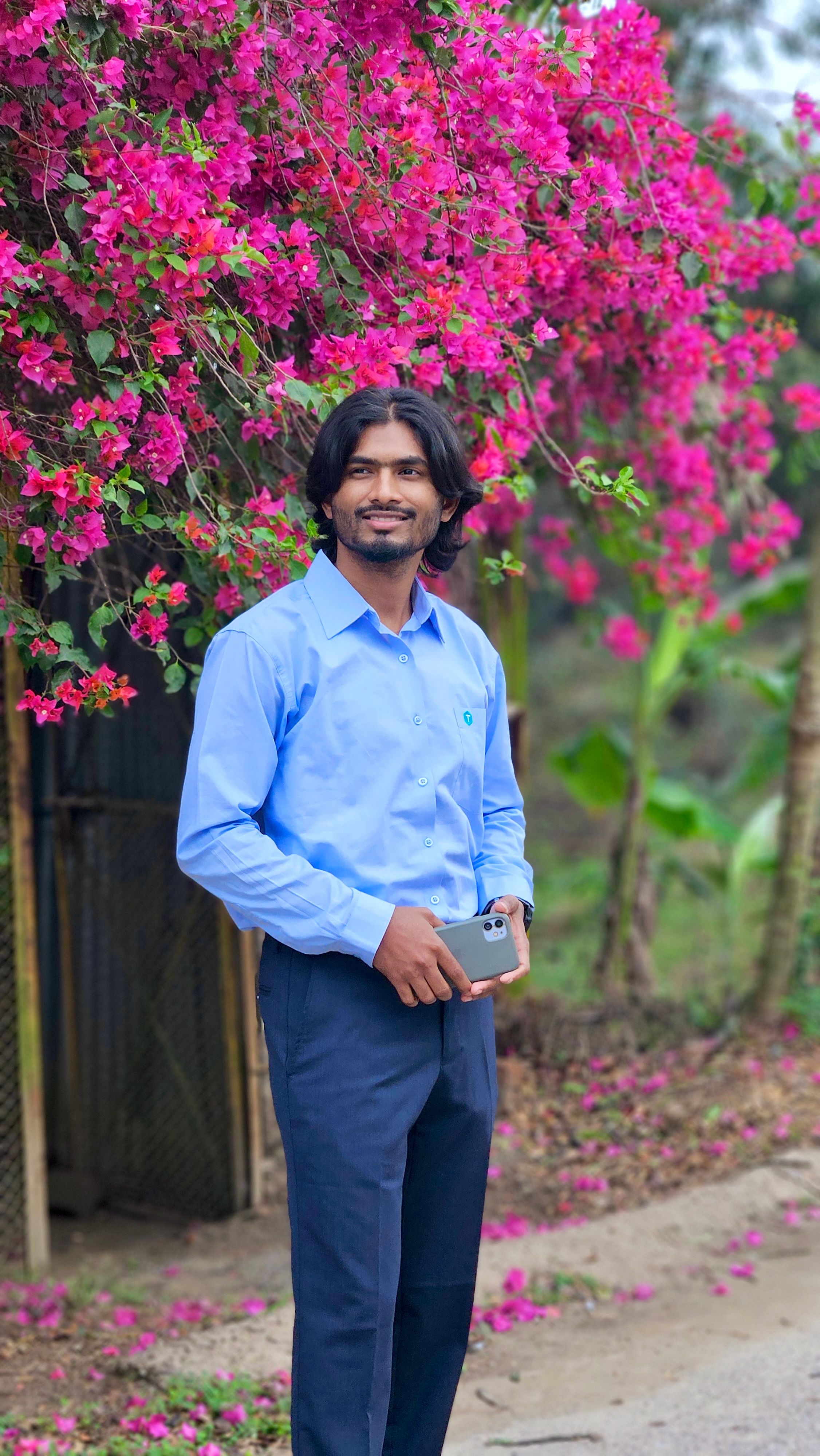 A smiling man in a blue shirt holding a phone, stands near blooming bougainvillea flowers outdoors.