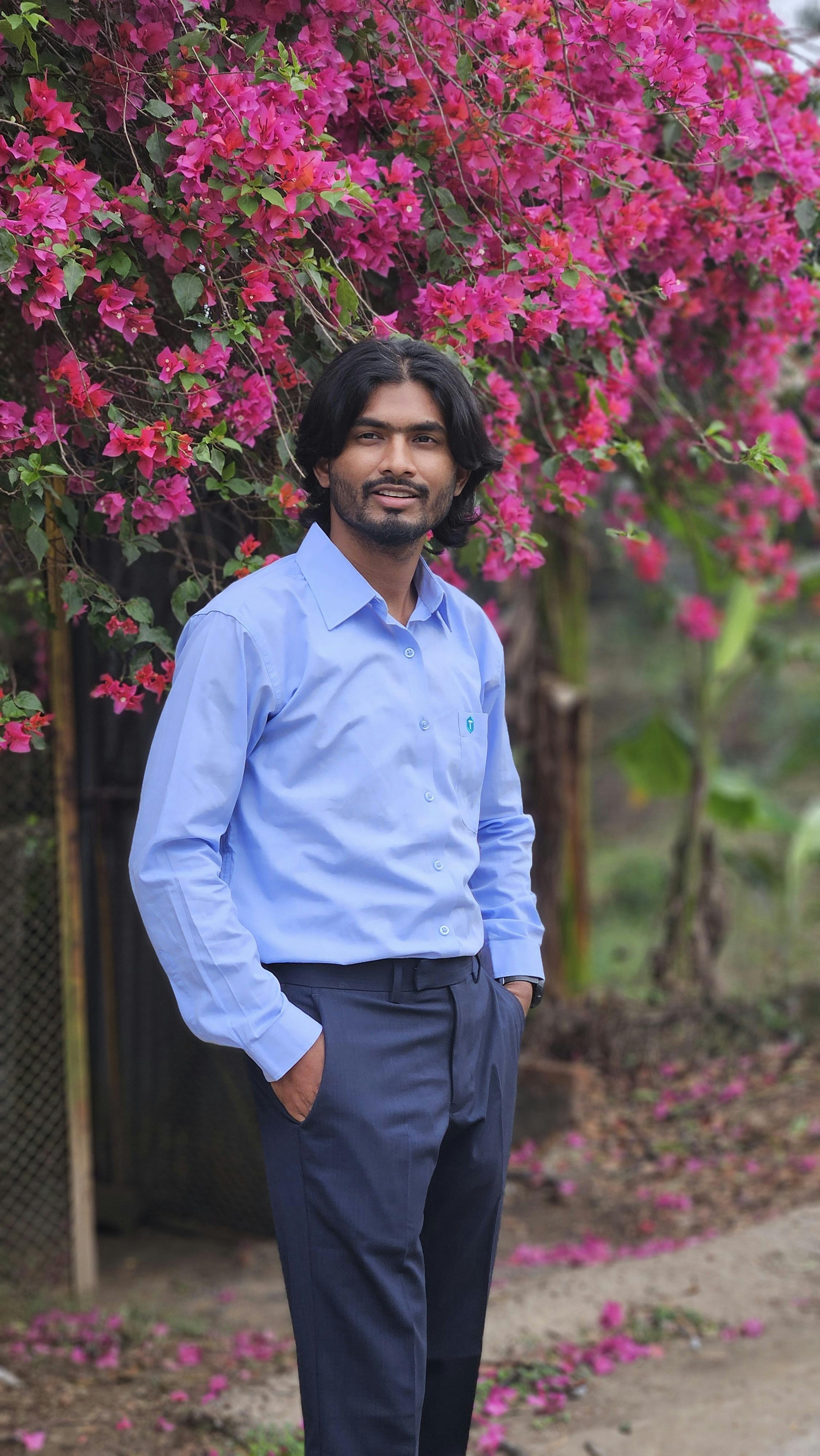 A young man in a blue shirt poses outdoors under a cascade of bright pink flowers.