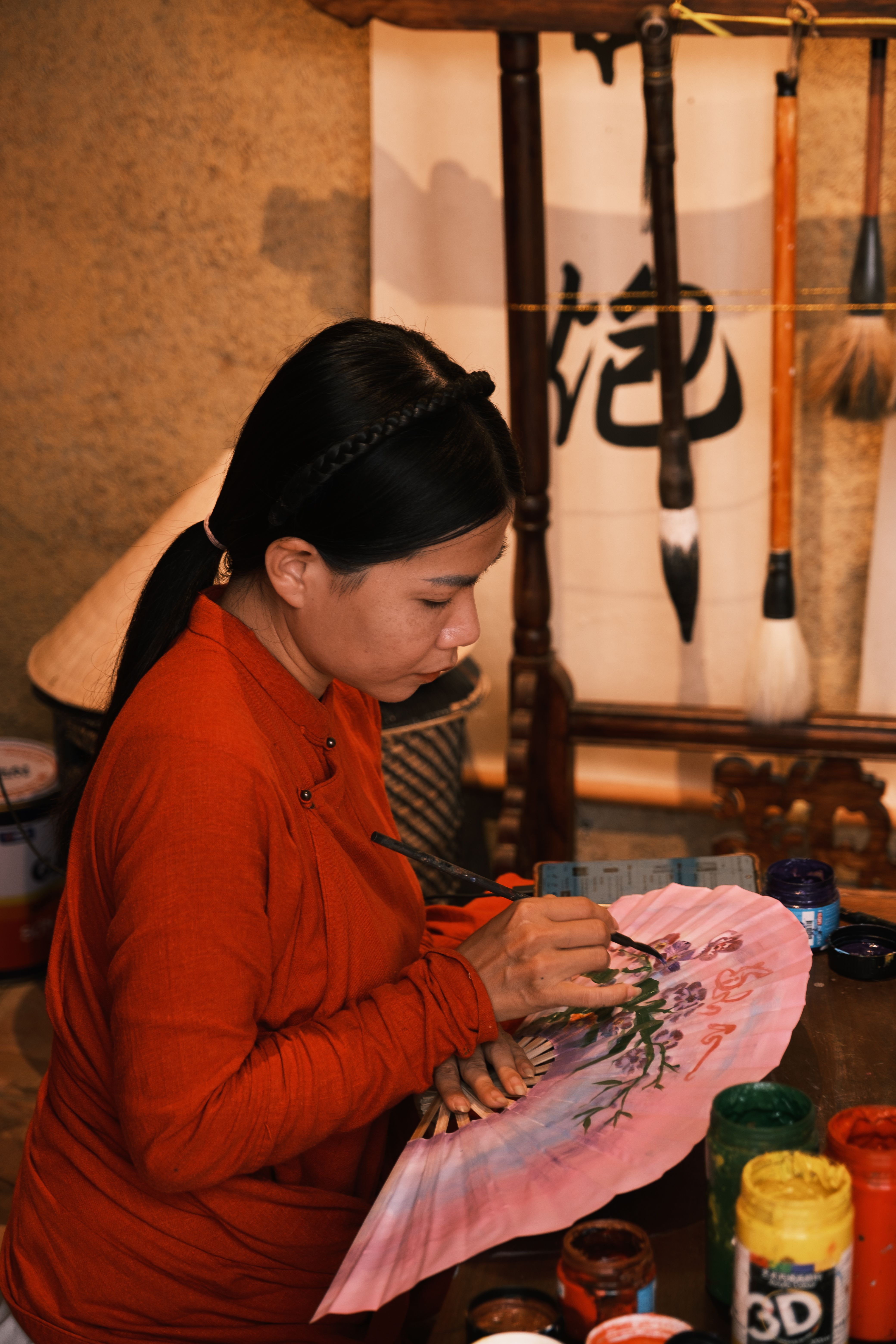 Woman artist creates a delicate flower design on a fan in a traditional Asian craft studio.