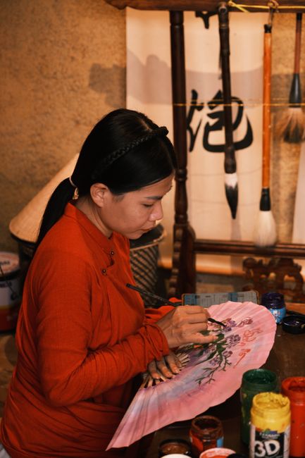 Woman artist creates a delicate flower design on a fan in a traditional Asian craft studio.