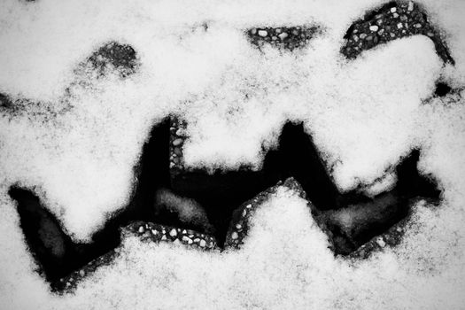 Black and white image of a crack in the snow revealing dark pebbles beneath, creating abstract patterns.