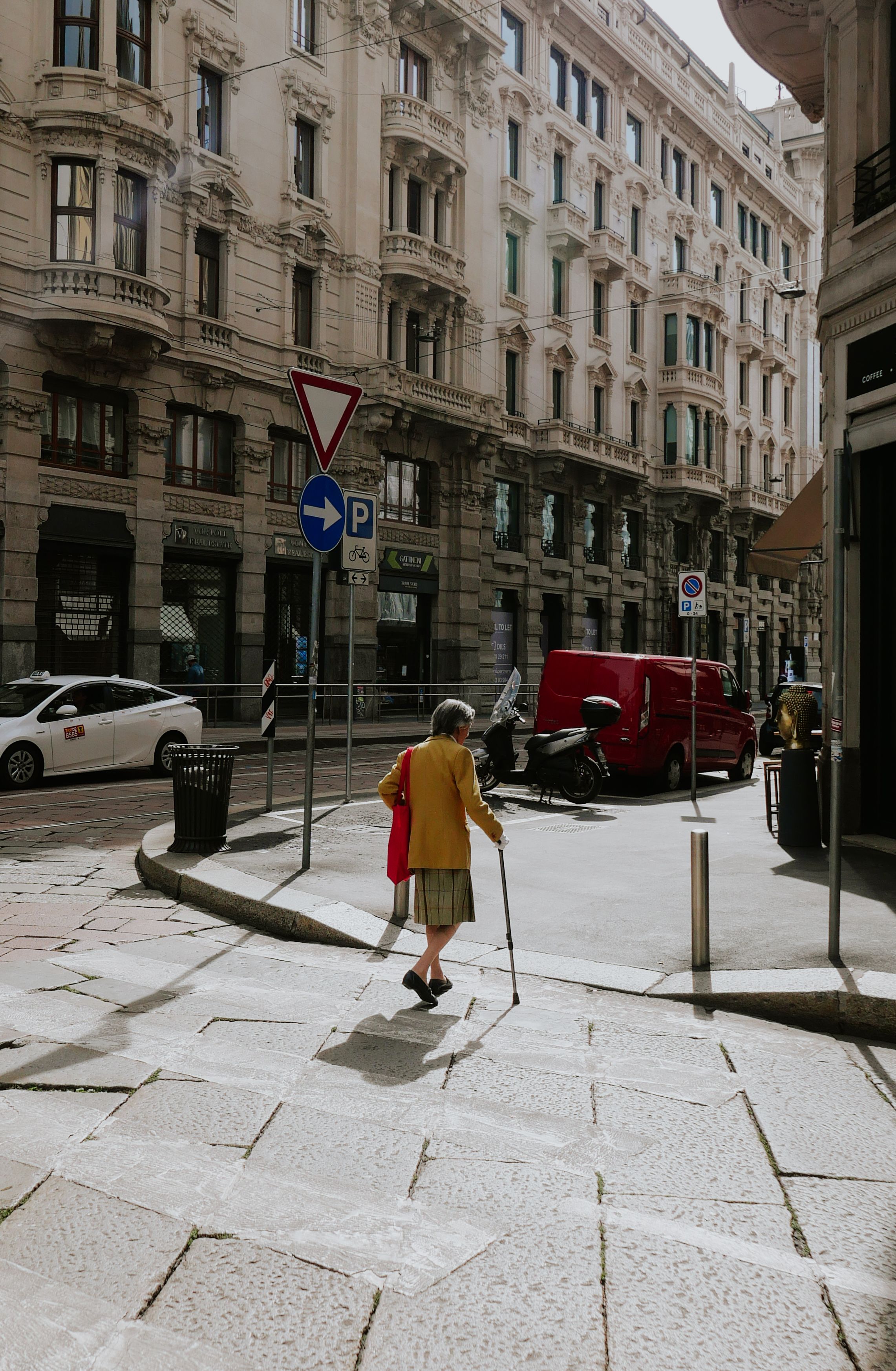 Elderly woman in yellow coat walking on a sunny day in Milan, Italy, surrounded by classic architecture.