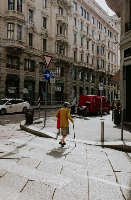 Elderly woman in yellow coat walking on a sunny day in Milan, Italy, surrounded by classic architecture.