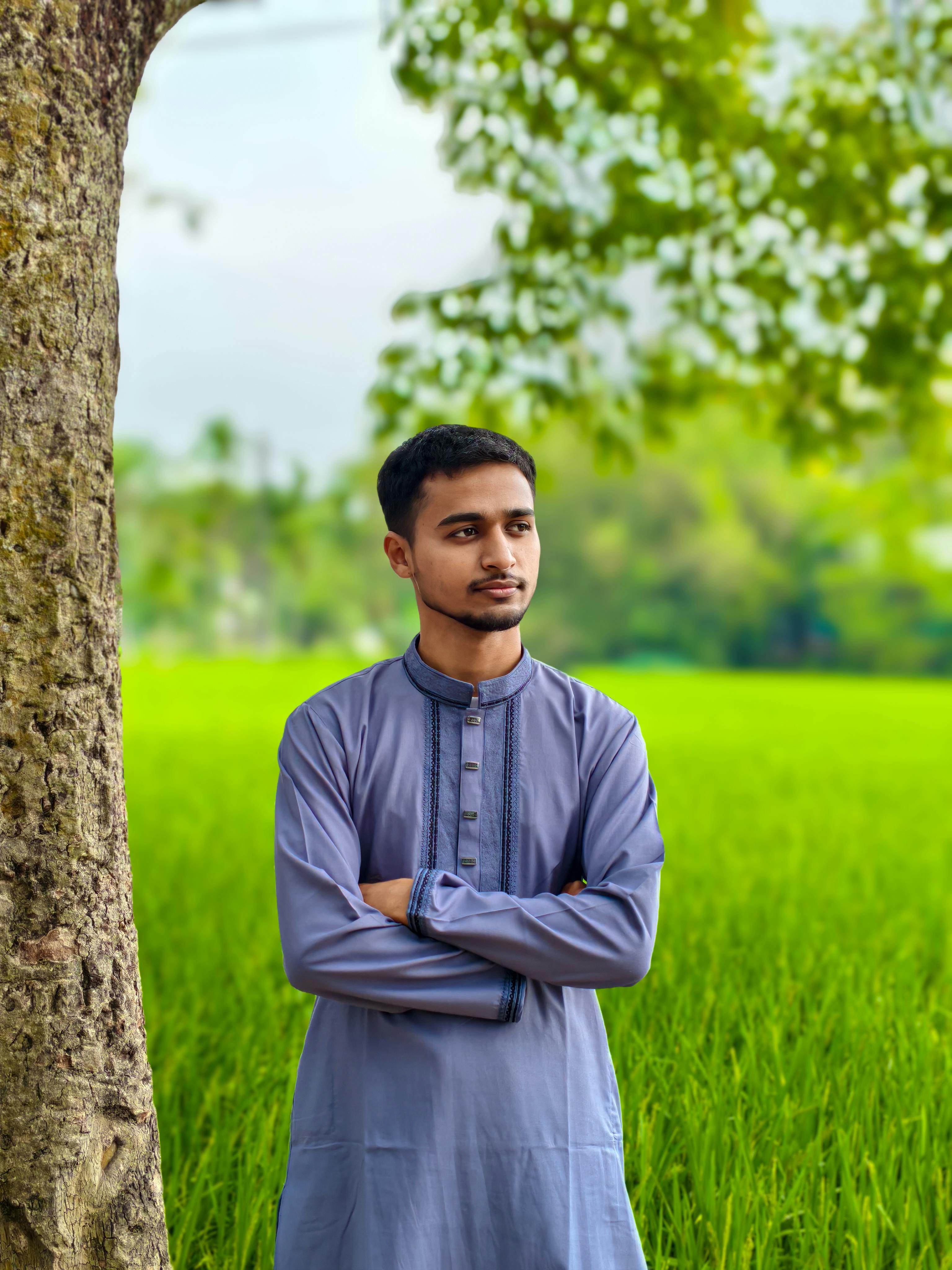 Young man in traditional clothing stands near a lush green field, framed by a tree, under a clear sky.