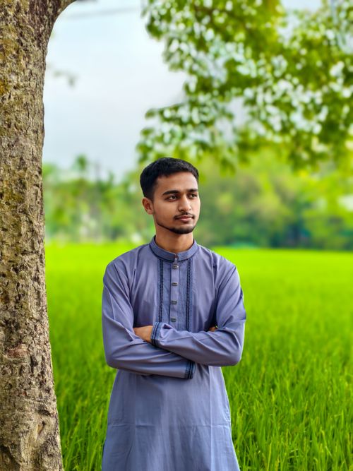 Free Young man in traditional clothing stands near a lush green field, framed by a tree, under a clear sky. Stock Photo