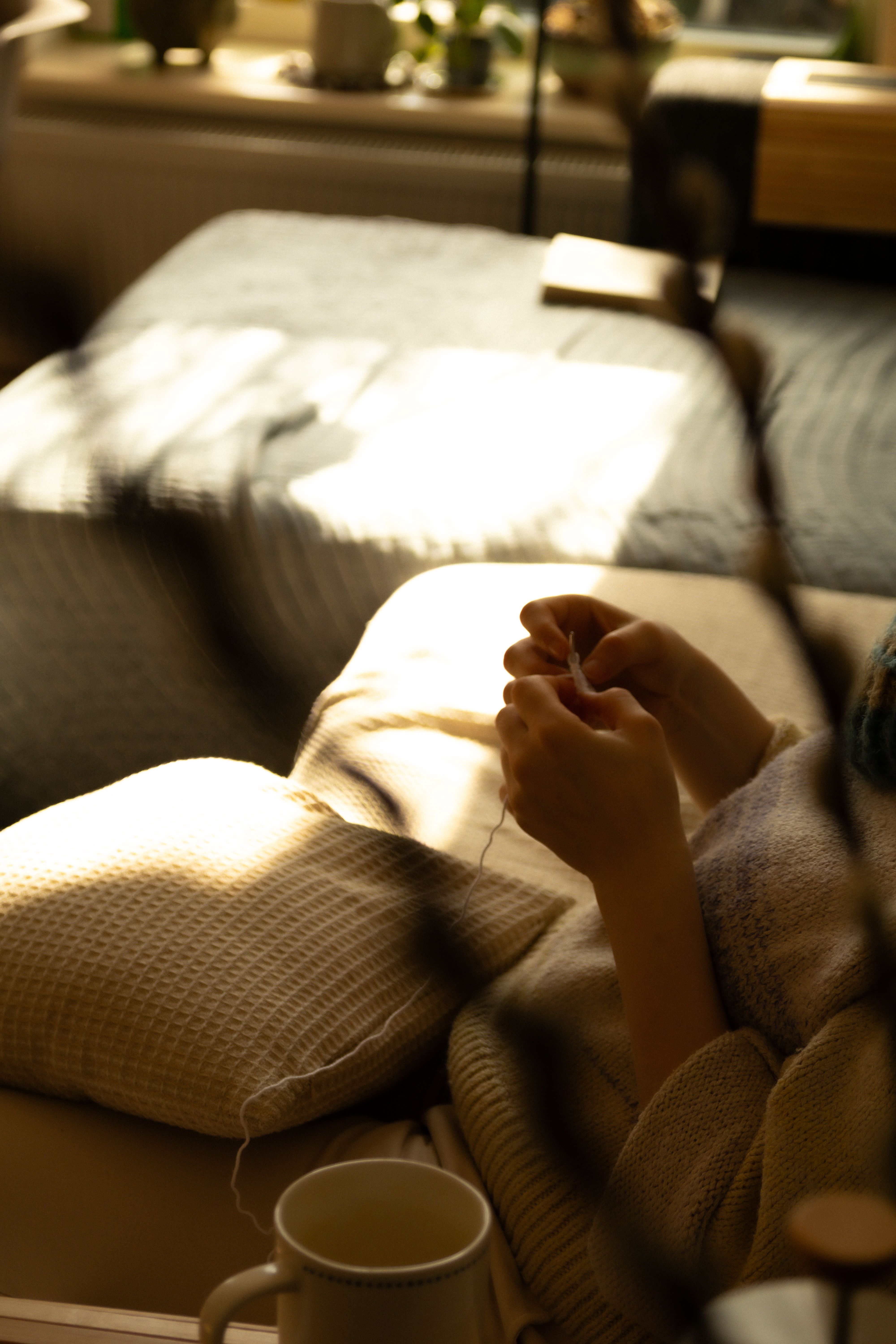 A person knitting indoors on a sofa, bathed in warm sunlight, creating a serene and relaxing atmosphere.