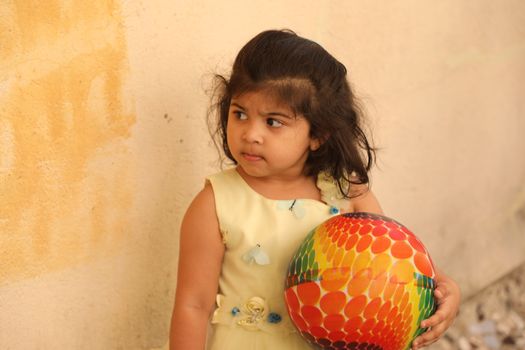Adorable child holding a colorful ball against a textured wall, looking curious.