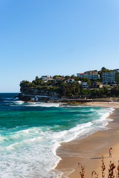 A vibrant coastal scene with waves, sandy beach, and homes perched on a cliff under a clear blue sky.