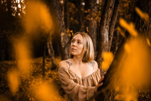 A woman in a brown dress poses in an autumn forest, surrounded by golden leaves and trees.