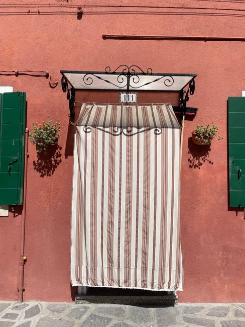 Charming Venetian doorway with a striped curtain on a rustic red wall and green shutters.