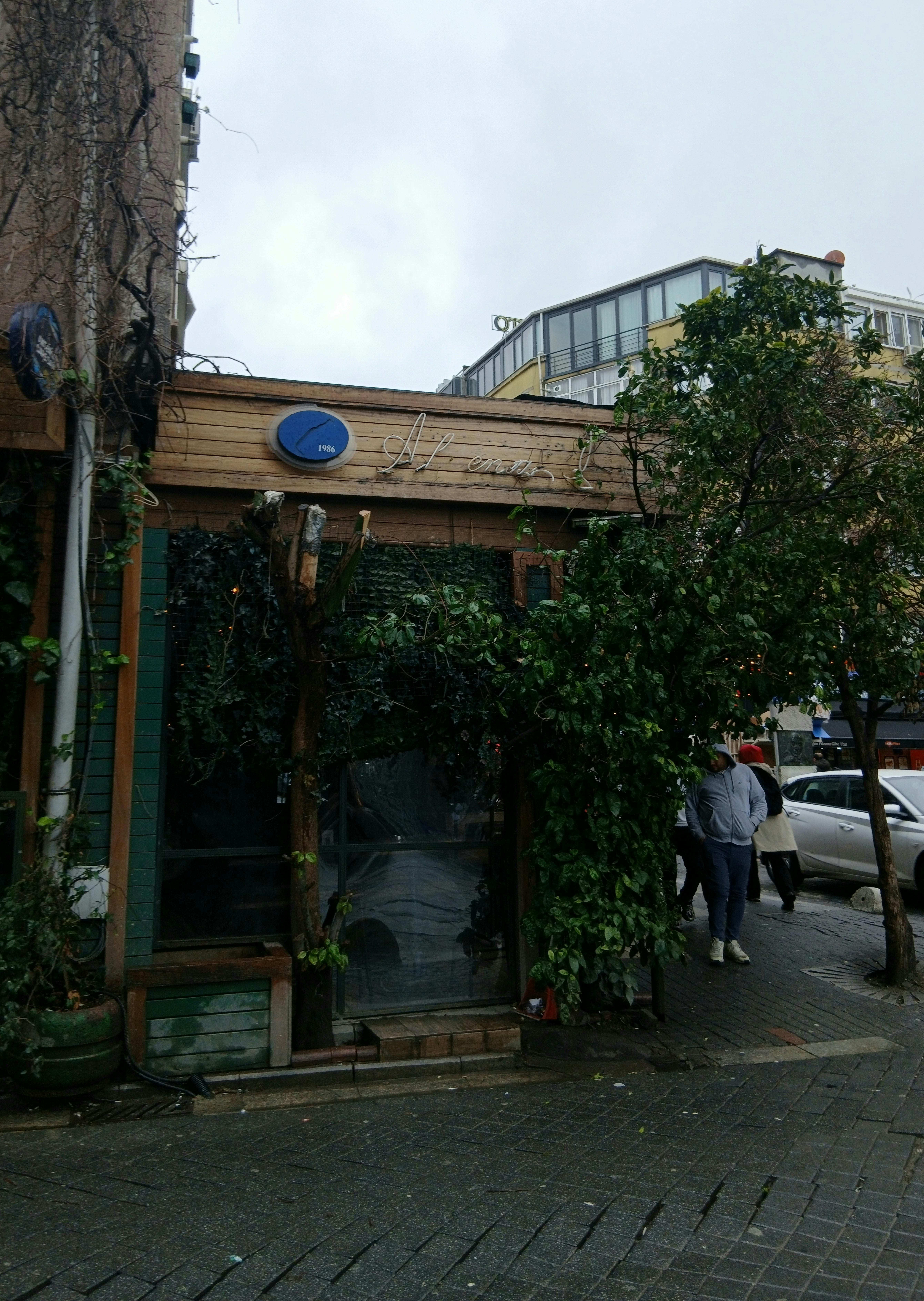 Street view of a cozy cafe with greenery on a cloudy day, featuring urban surroundings.