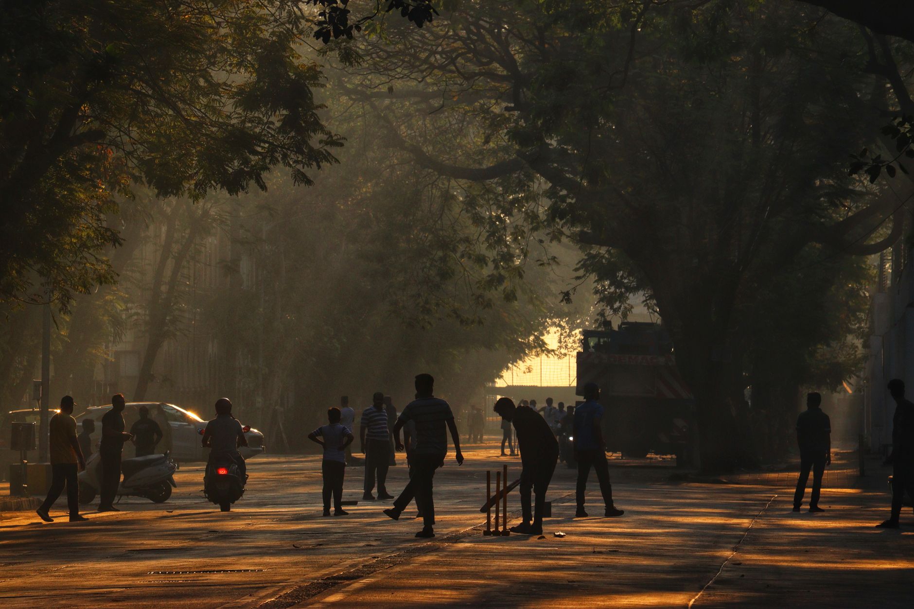 Silhouetted people playing cricket on a tree-lined street in Mumbai at dawn.