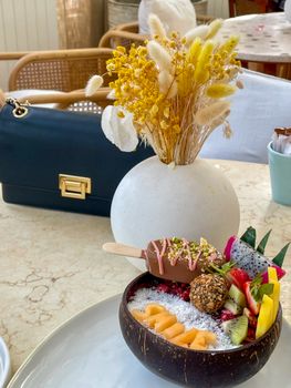 Colorful fruit bowl with dried flowers and a stylish handbag on a marble table.