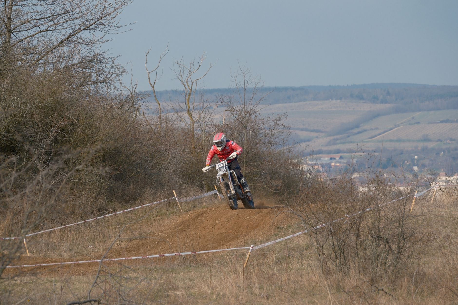 A motorcyclist competes in an off-road race in Jihomoravský kraj, Česko, surrounded by countryside landscape.