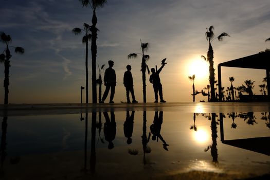 Three silhouetted people with palm trees reflected in water during a sunset.