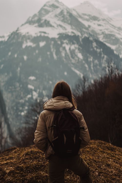 Free Traveler with backpack enjoying majestic snow-capped mountain views on a cloudy day. Stock Photo