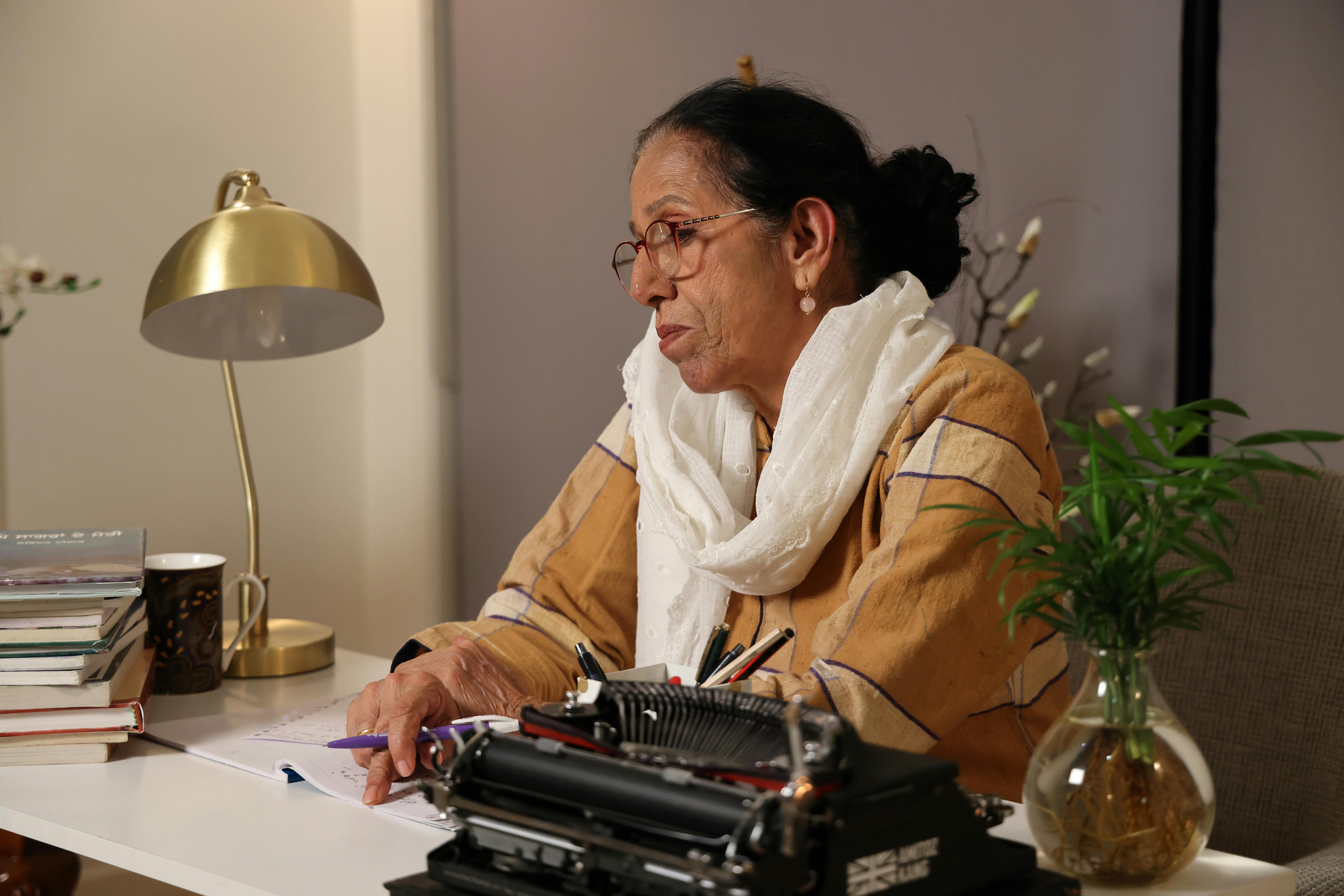 An elderly woman sits at her desk writing with a typewriter in a cozy indoor setting.