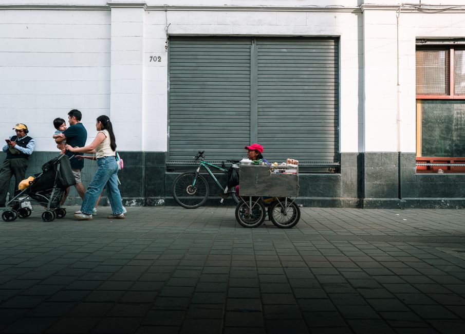 Vista de una calle urbana con familias, un vendedor y un local comercial cerrado.