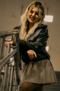 A fashionable woman poses on an indoor stairway, wearing a leather jacket and skirt.