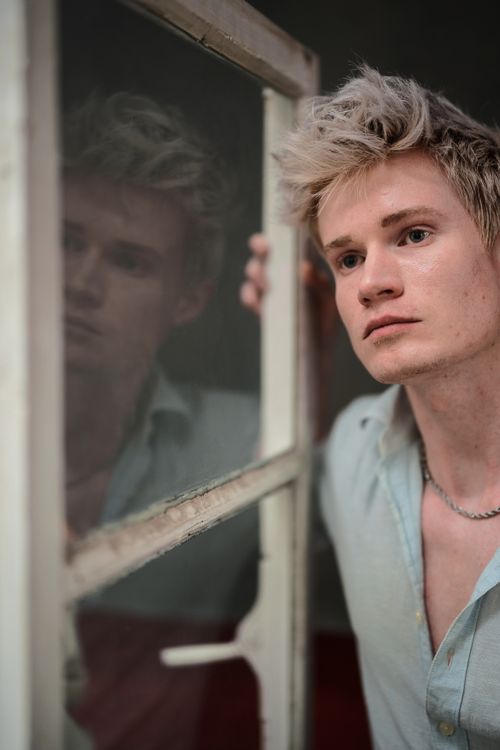 Free Thoughtful young man standing by a window, gazing outside. Moody and reflective atmosphere. Stock Photo