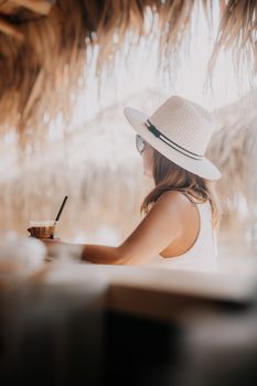 A woman in white sunhat relaxing with an iced coffee under a beach hut.