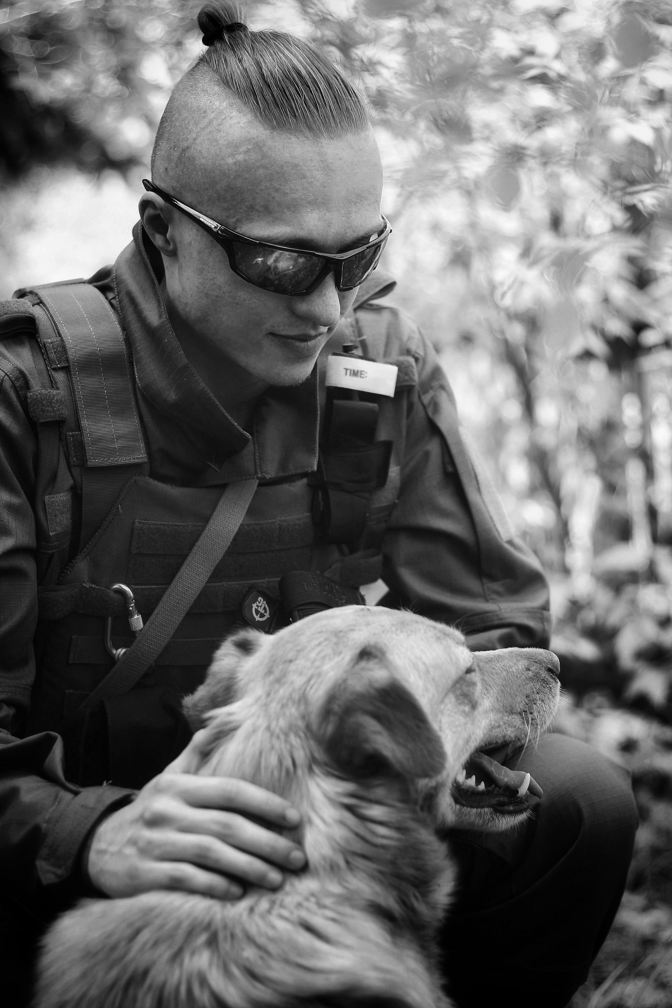 A soldier wearing tactical gear interacts with a dog in a wooded area, showing companionship.