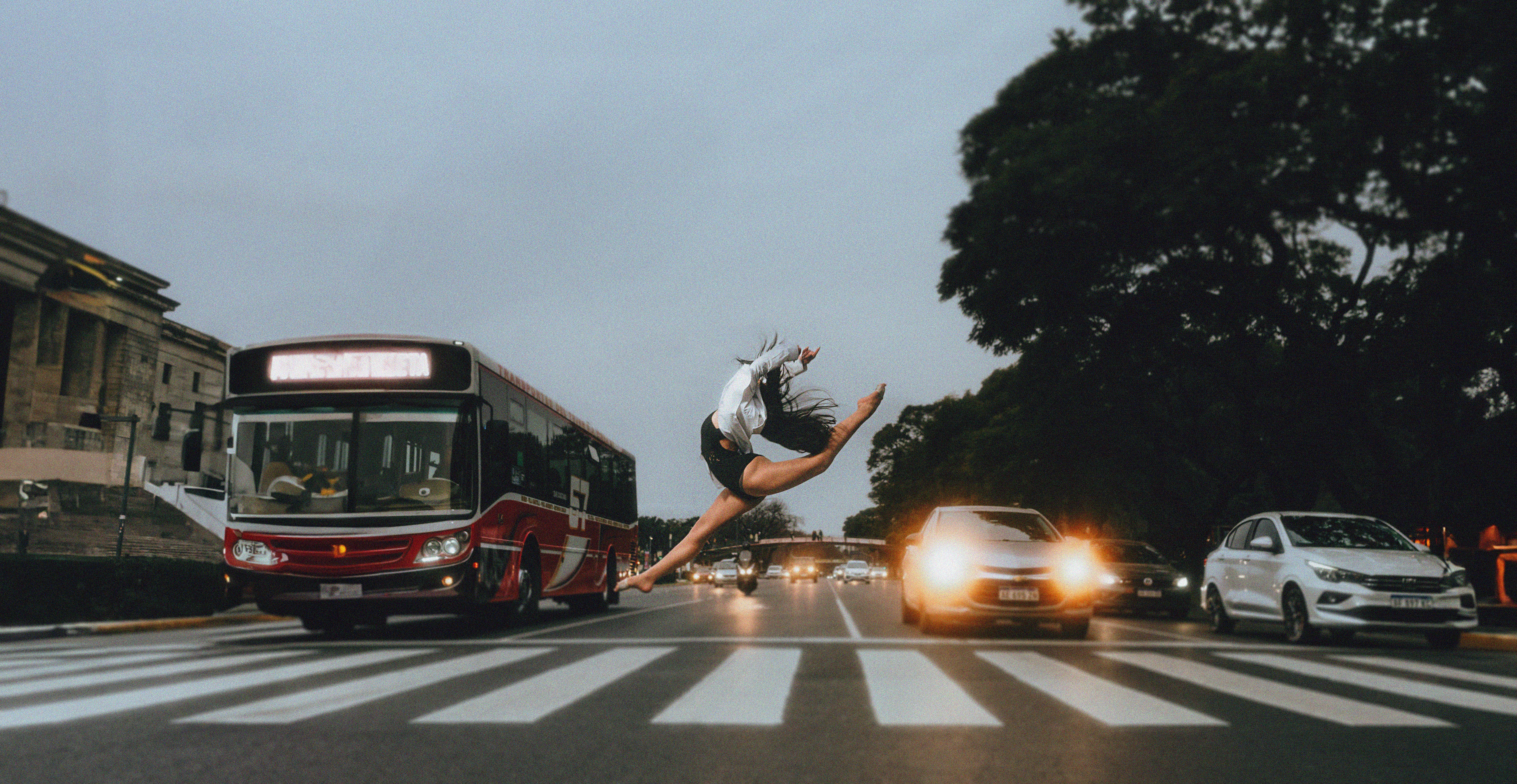 A dancer dramatically leaps over a zebra crossing amidst city traffic at dusk.