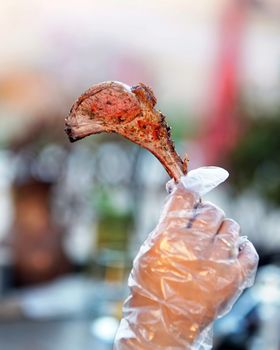 A deliciously seasoned grilled lamb chop held vertically in a gloved hand, set against a blurred background.