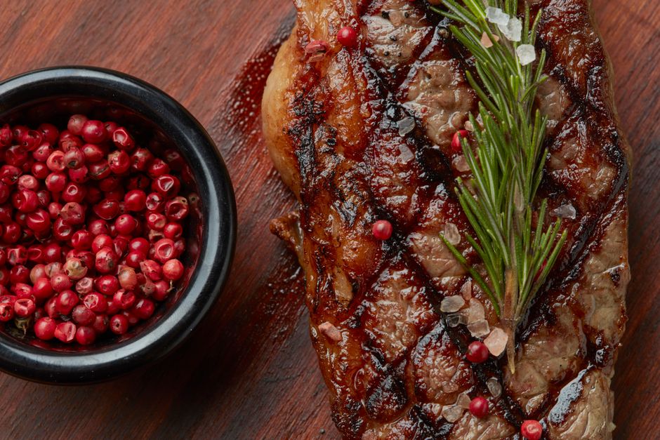 A small wooden bowl of coarse steak seasoning with visible flecks of garlic, black pepper, and red pepper next to raw steaks on a cutting board