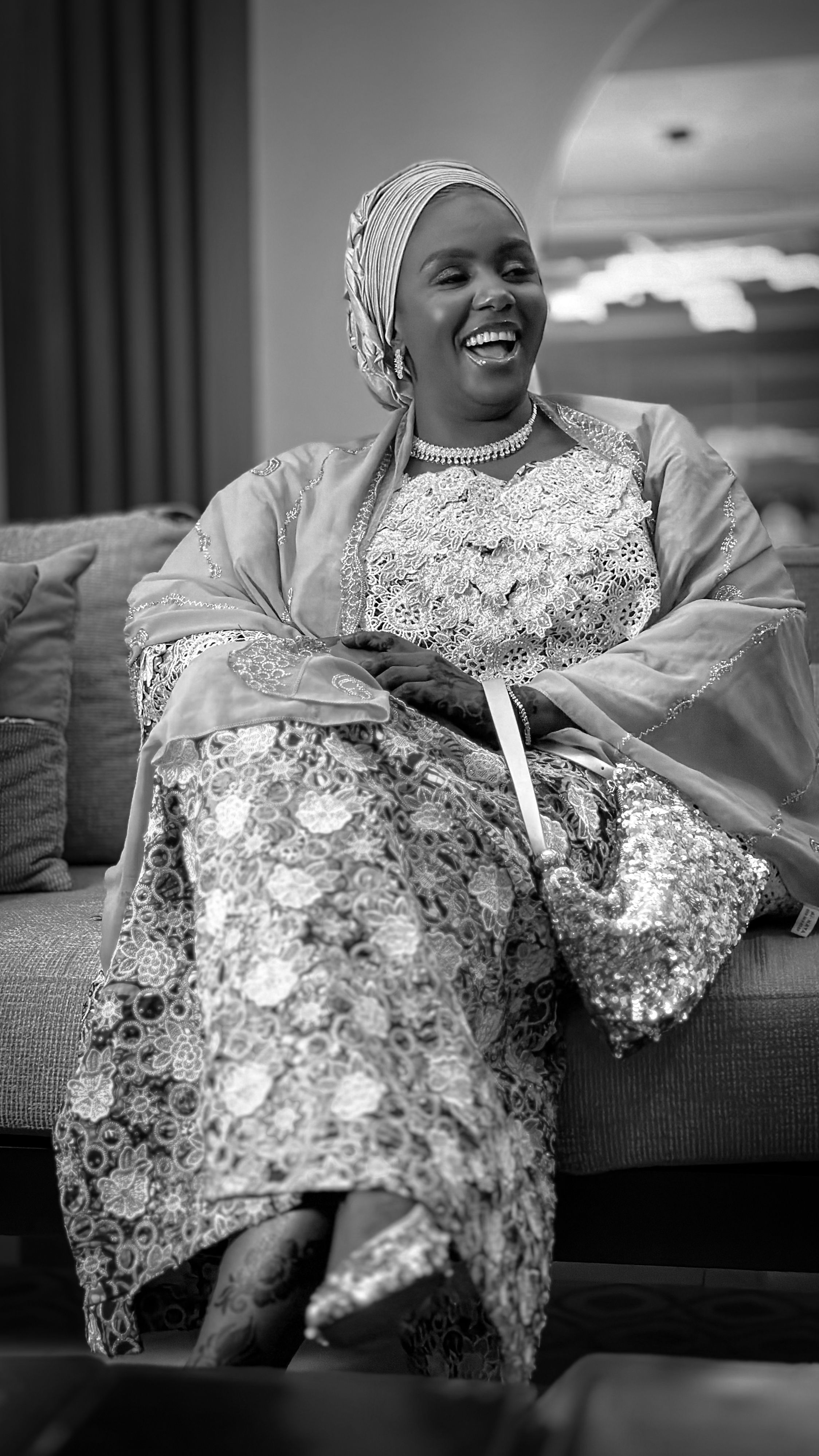 Smiling woman in intricate African attire seated indoors, exuding joy and elegance.