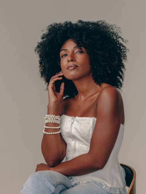 Free Elegant portrait of a woman sitting in a white top and jeans, showcasing natural curly hair and pearl jewelry. Stock Photo