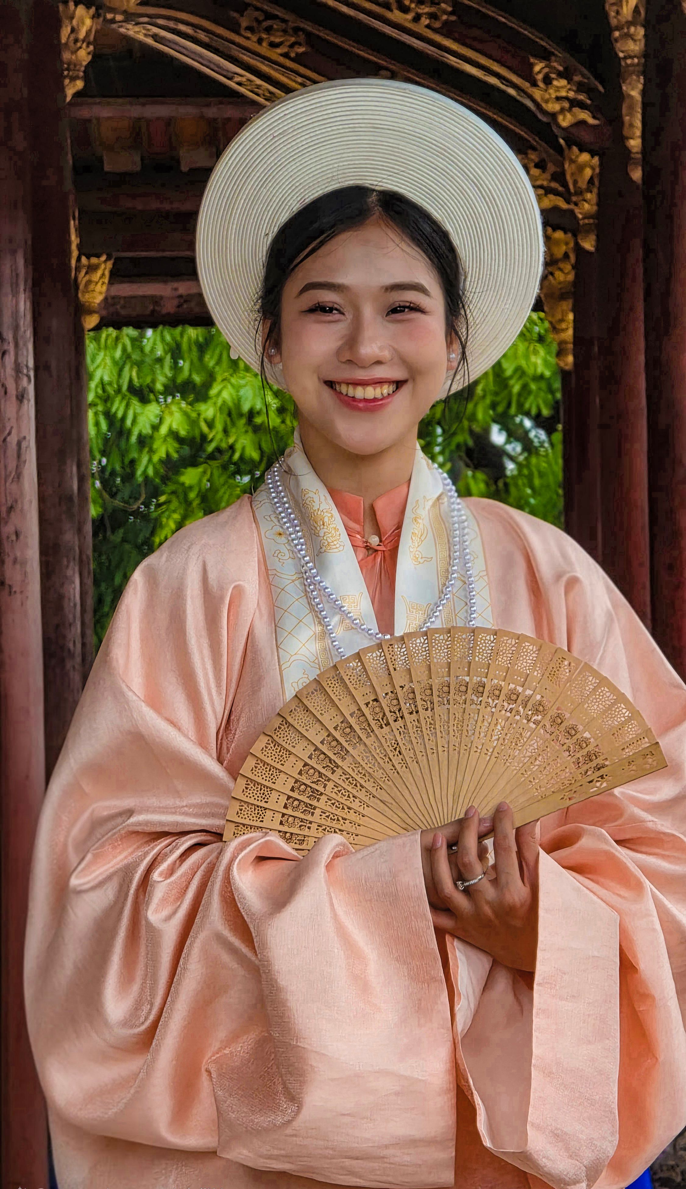 Smiling young woman in Vietnamese traditional dress holding a fan at a cultural landmark.
