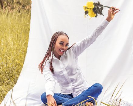 A cheerful woman sitting on grass holding yellow flowers against a white backdrop, enjoying a sunny day.