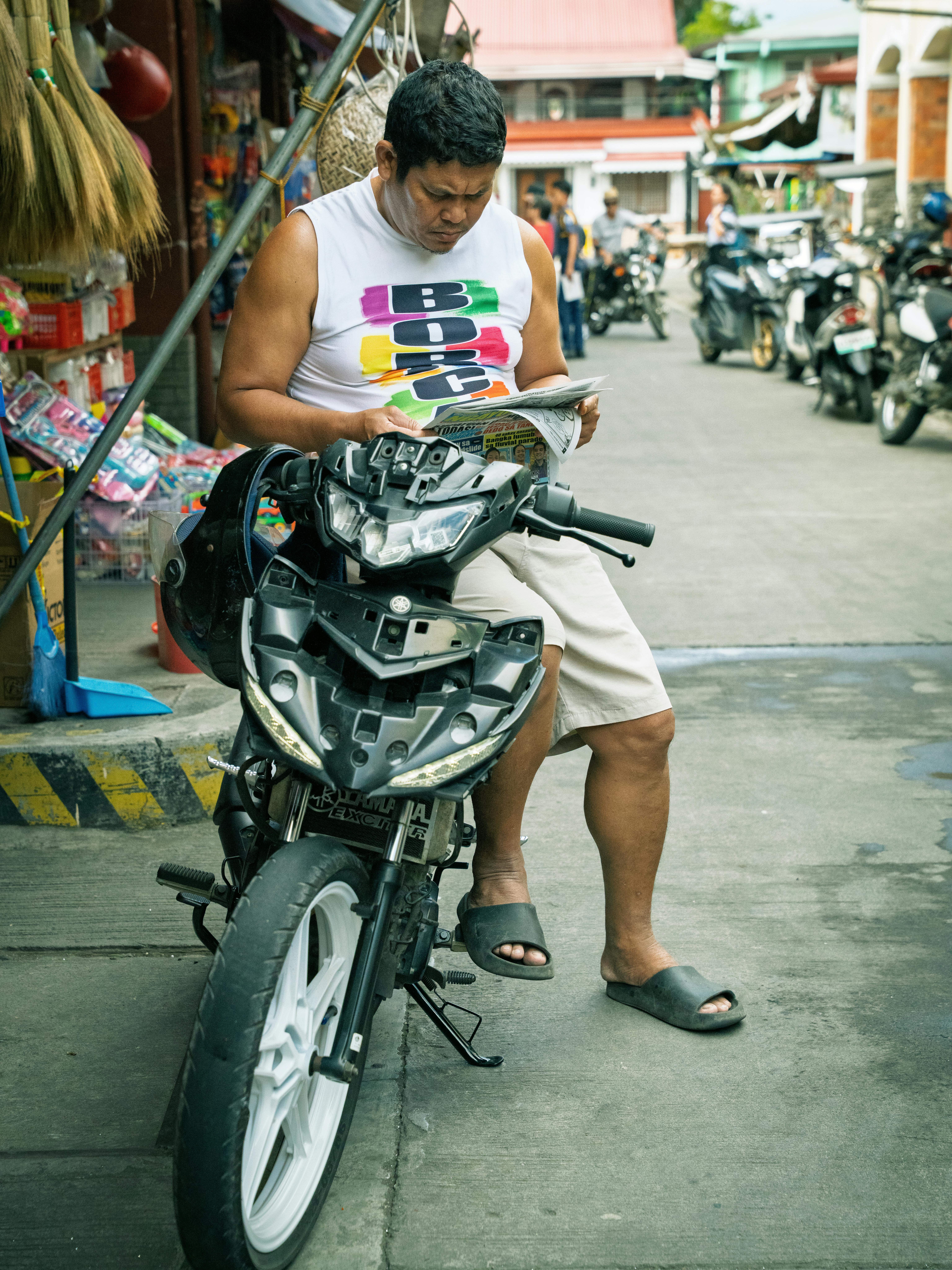 A man casually reading a newspaper while seated on a motorcycle in the vibrant streets of Taal, Philippines.