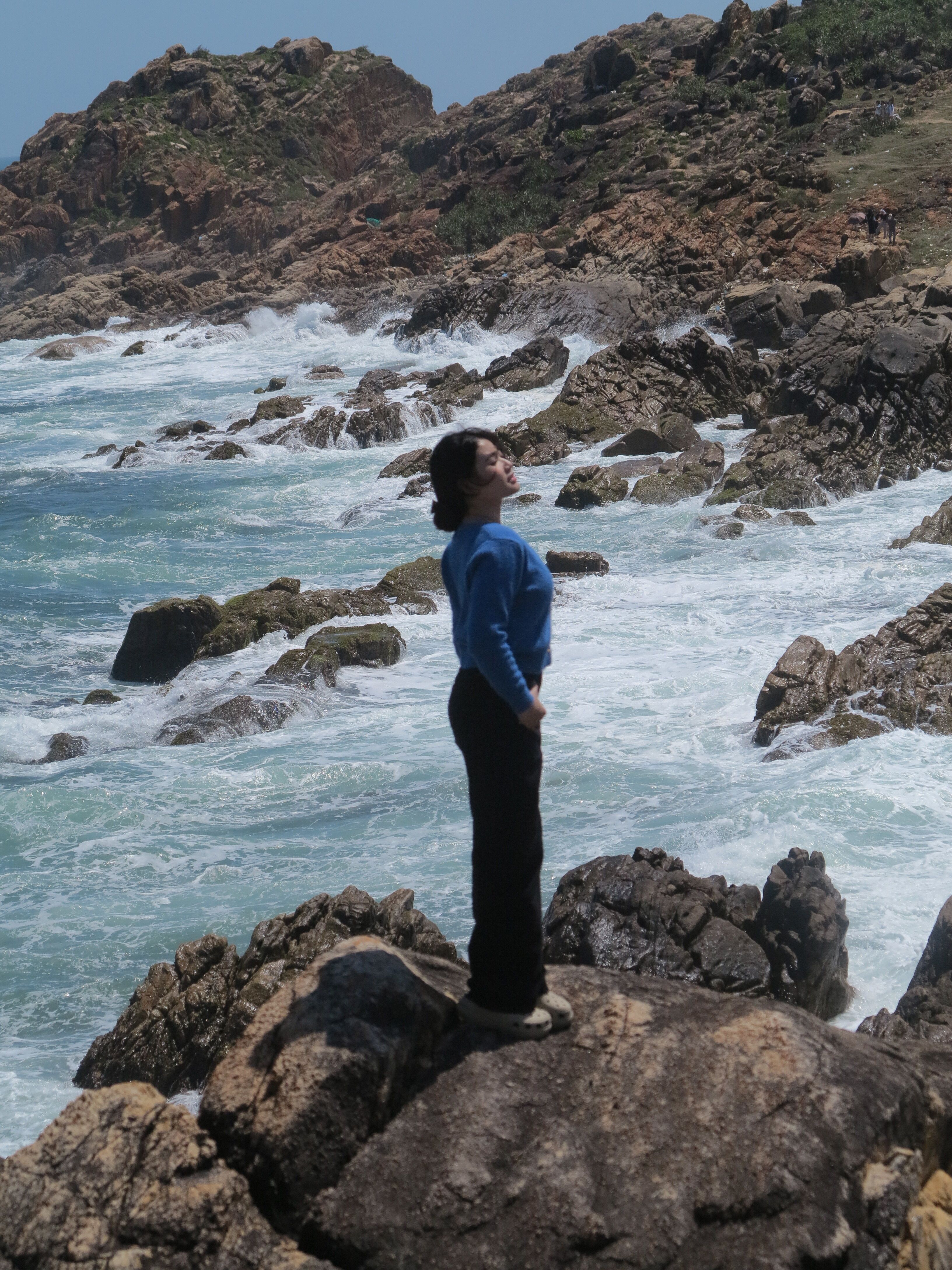 A woman in a blue sweater stands on a rocky shore near the ocean waves at Phú Yên, Vietnam.