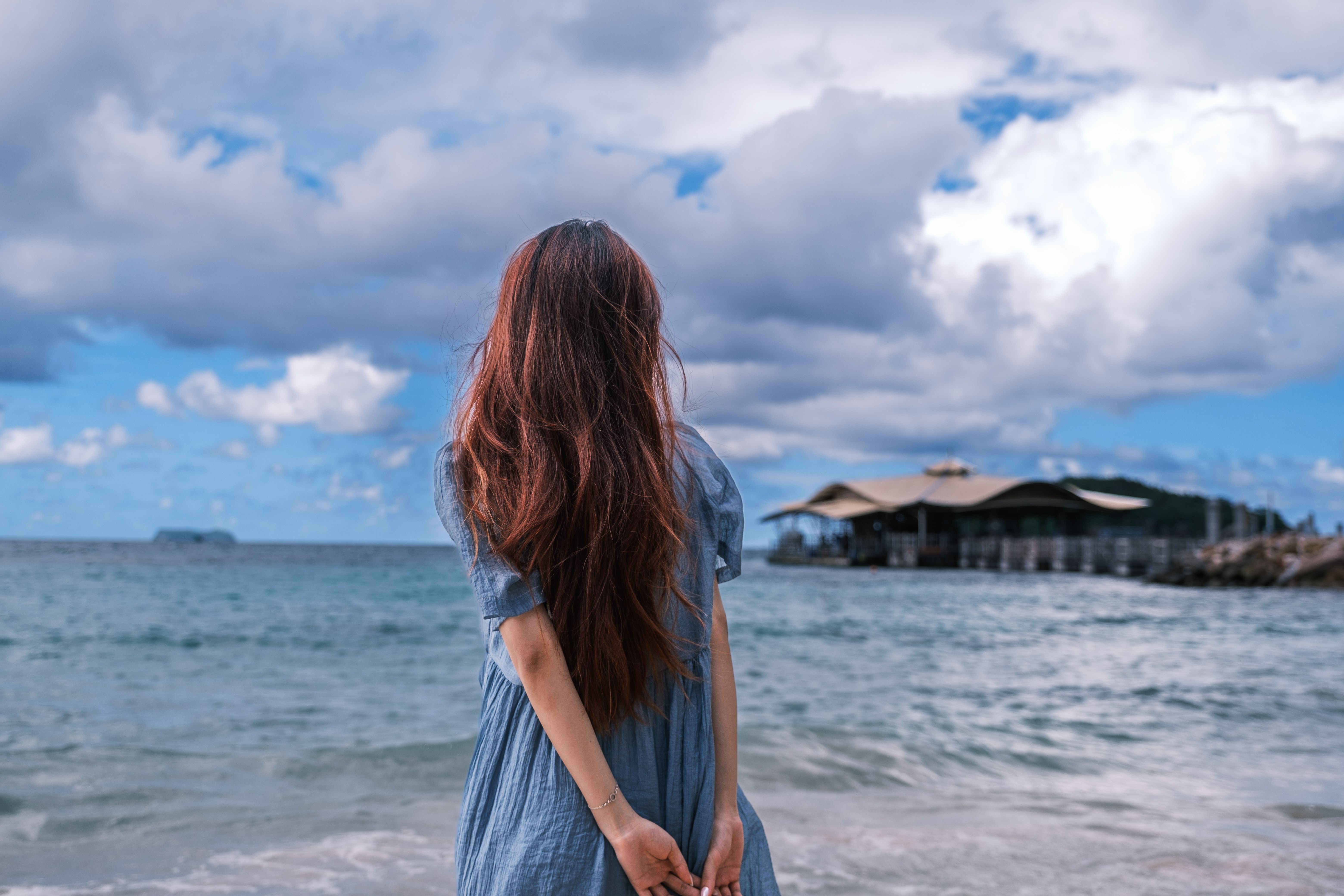 A young woman with long hair stands on a beach, looking at a beautiful ocean view under a cloudy sky.