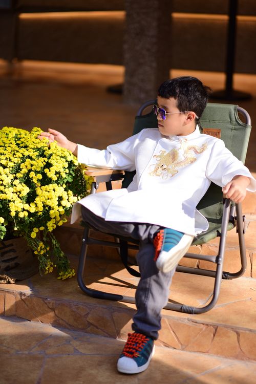Free Young boy in sunglasses and traditional attire, seated outdoors beside vibrant yellow flowers. Stock Photo