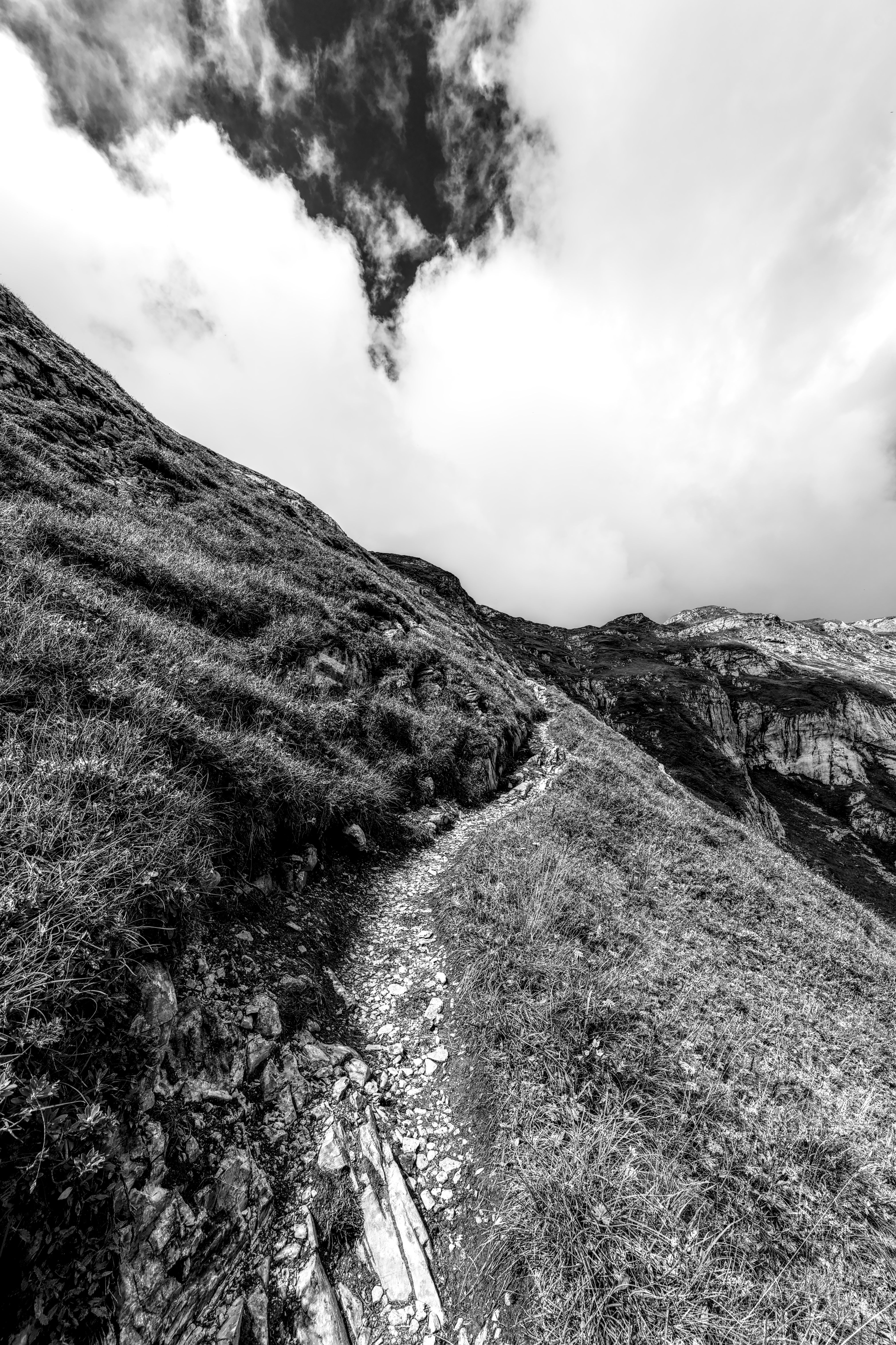 Dramatic black and white photo of a rugged mountain trail under a cloudy sky.