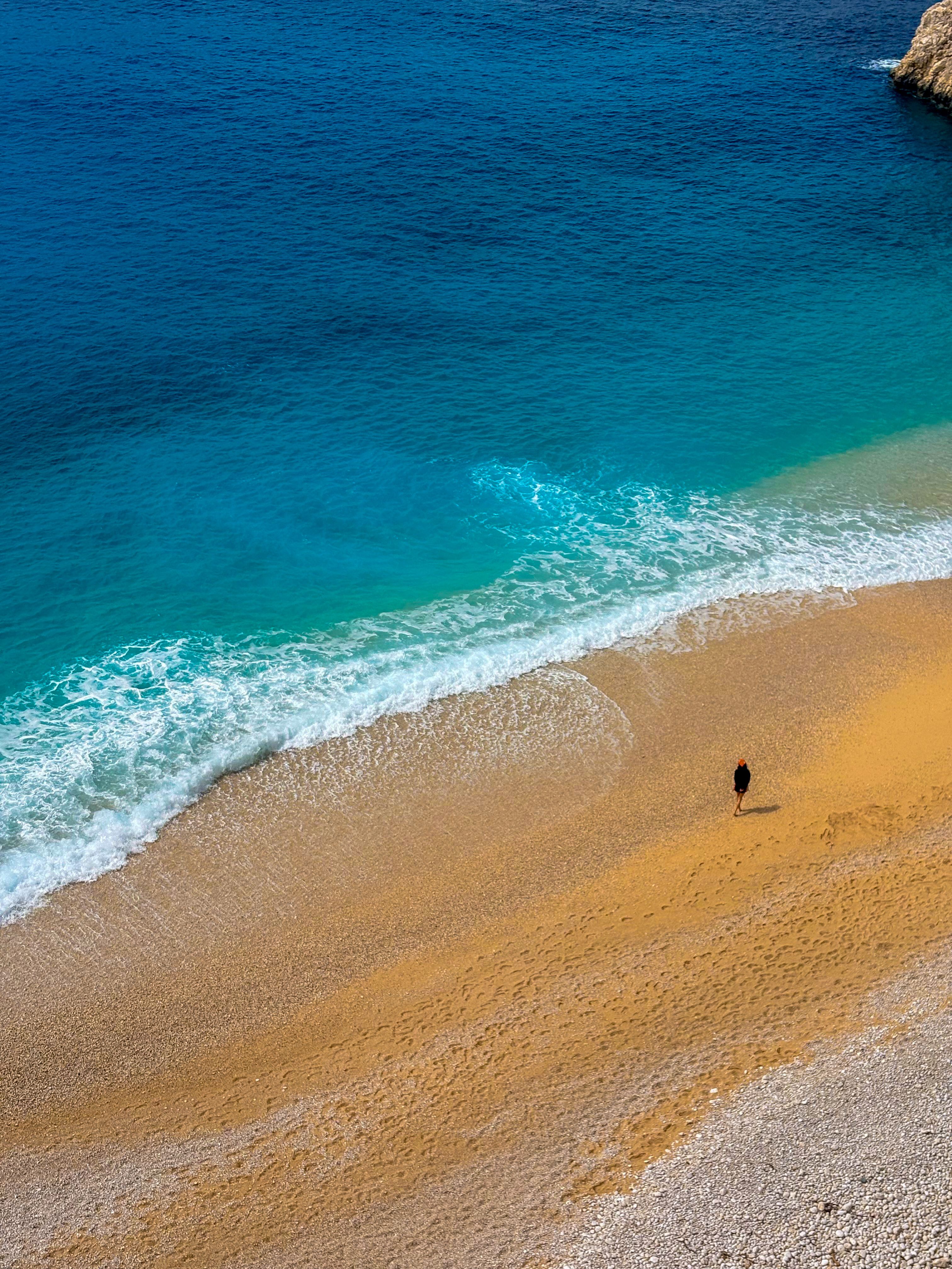 A lone figure walks along the pristine beach of Kaş, Antalya, Türkiye, under a bright day sky.