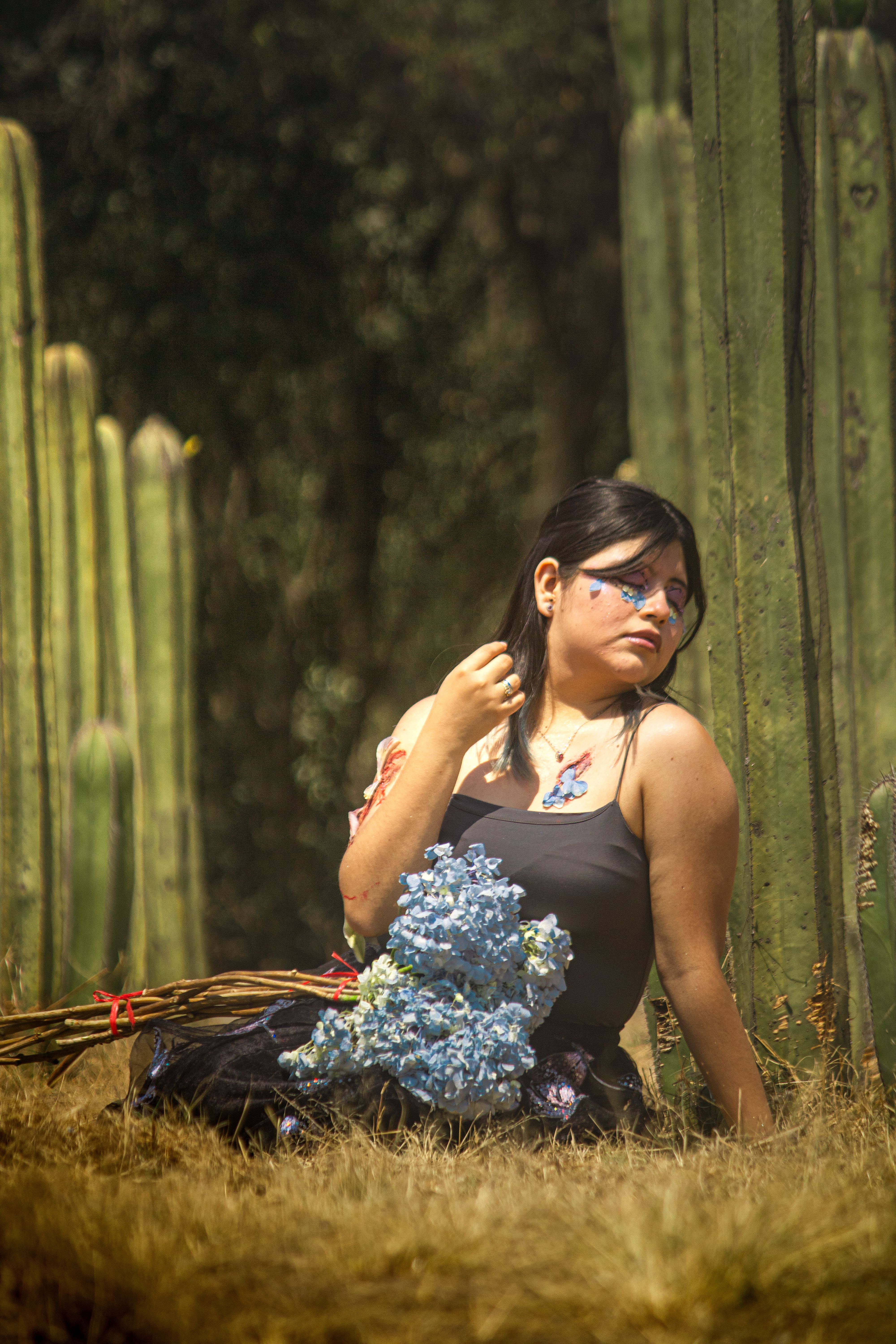 A woman with artistic makeup poses among cacti in a sunlit garden, holding a bouquet of blue flowers.