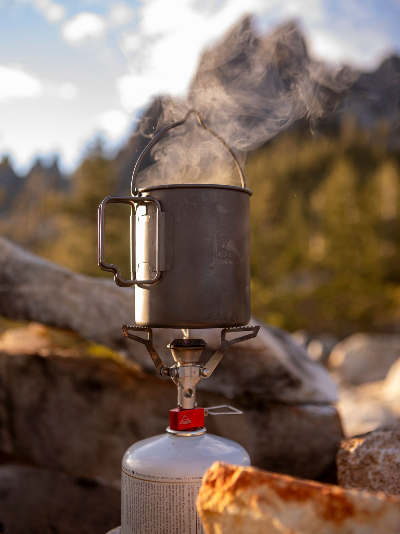Camping stove heating a metal pot among rocks in a mountain landscape