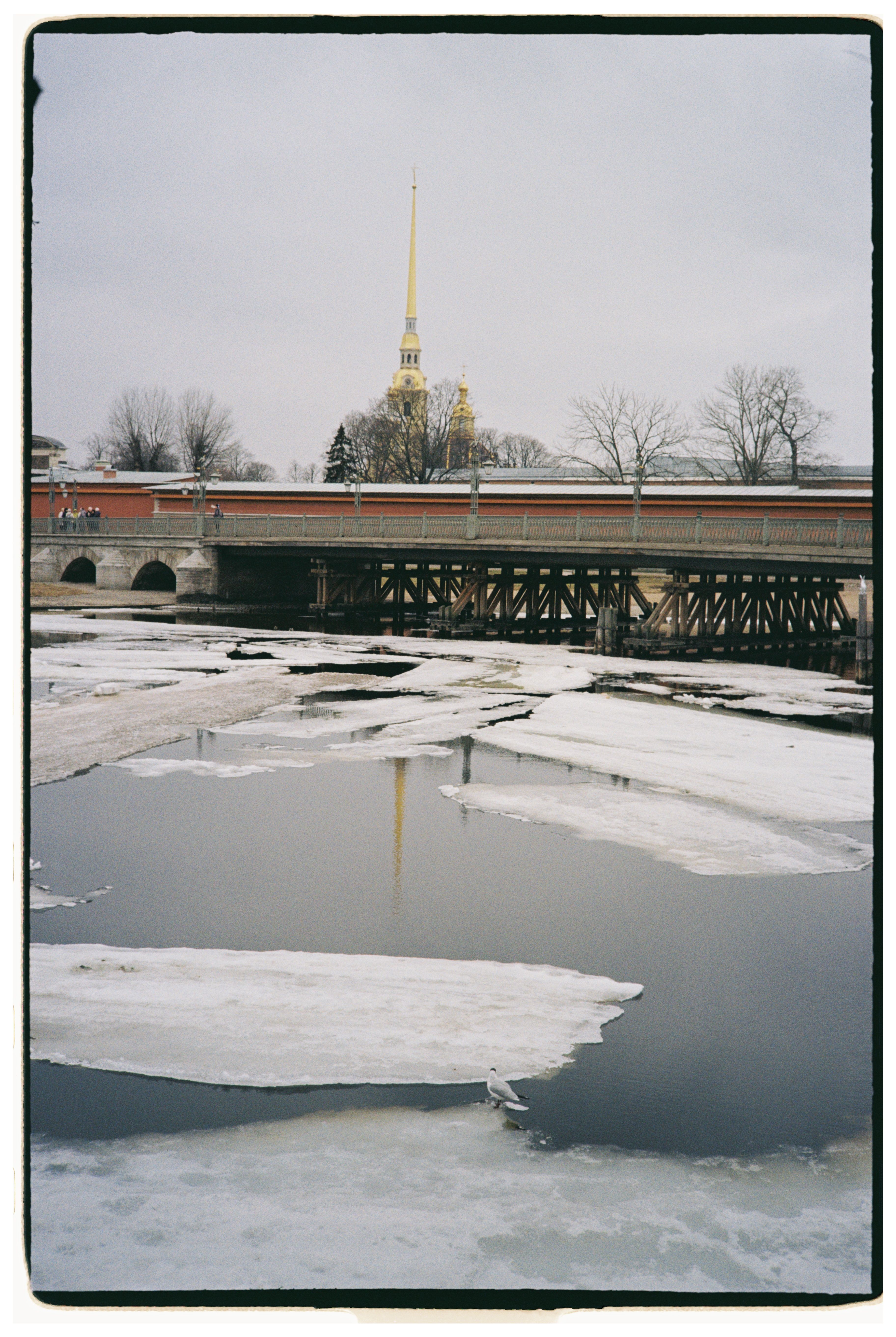 A winter scene of a frozen river and a bridge with a distant pointed spire under cloudy skies.