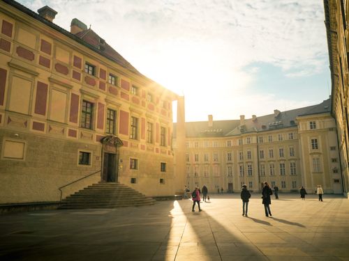 Free A sunlit courtyard featuring historical European architecture and people walking, casting long shadows. Stock Photo