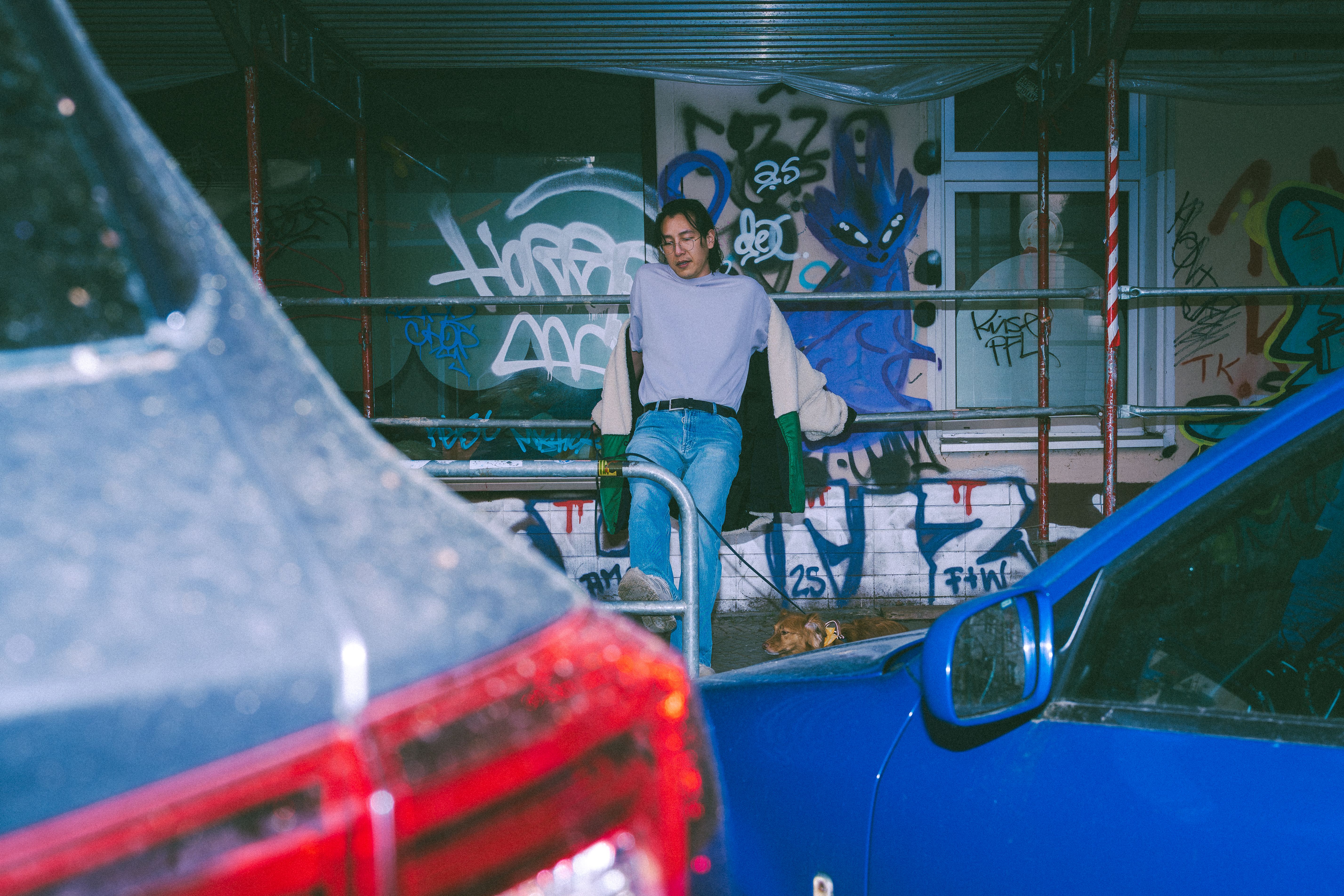 Jovem adulto posando perto de um muro grafitado em Berlim, cercado por carros estacionados.